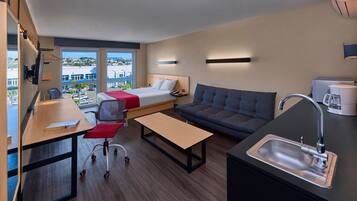 A modern hotel room with a bed, sofa, desk, and kitchenette.