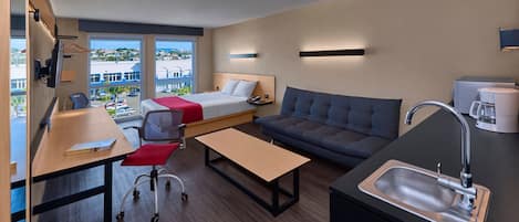 A modern hotel room with a bed, sofa, desk, and kitchenette.