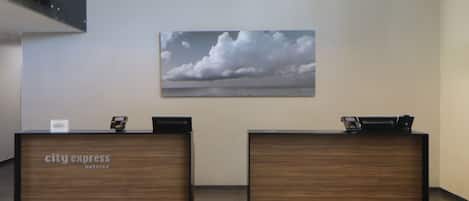 Un comptoir d’accueil avec une enseigne « City Express Hotels », deux postes de travail pour les réceptionnistes et un grand tableau représentant un paysage nuageux accroché au mur.