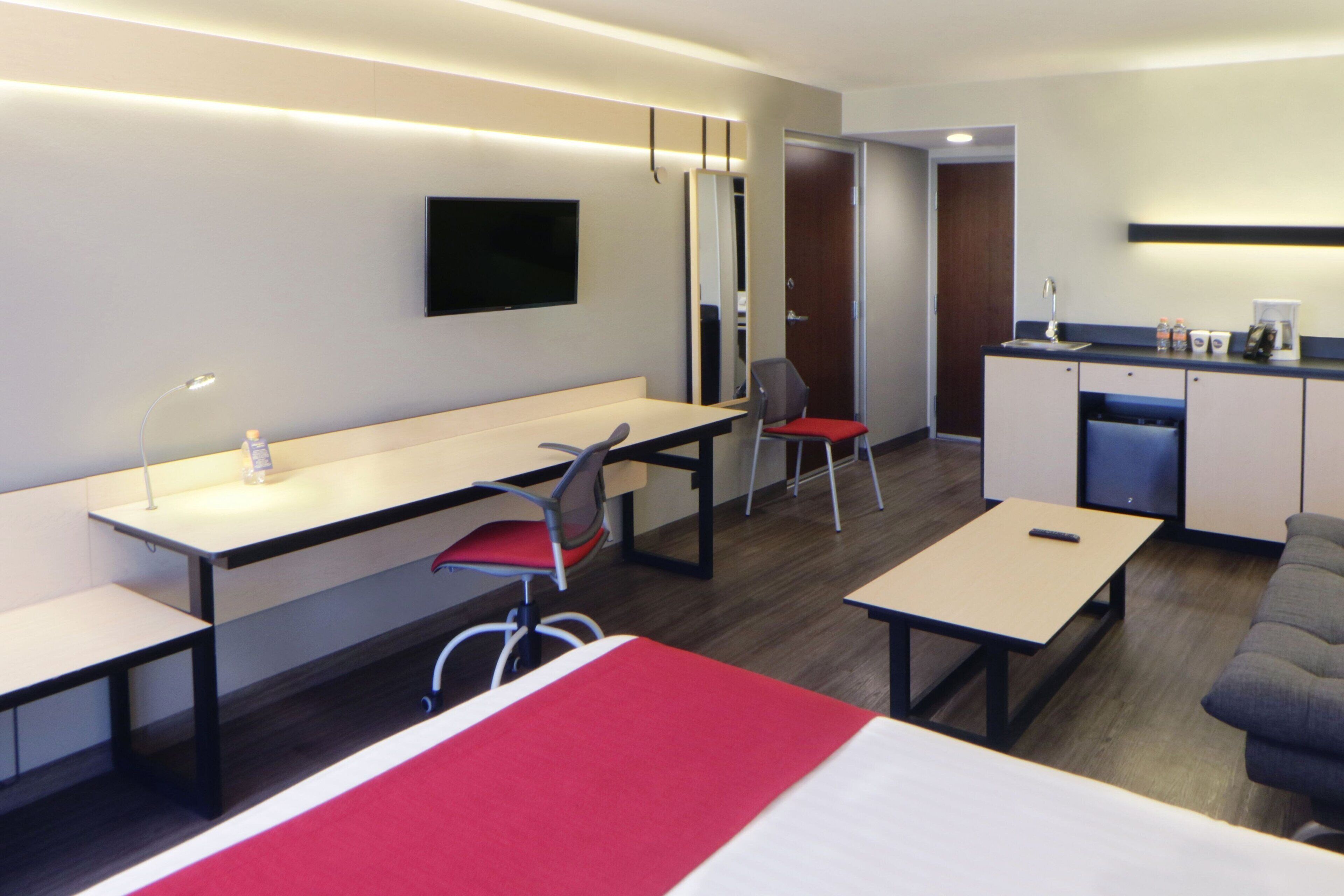 A modern hotel room with a bed, a desk, a chair, a sofa, a TV, and a kitchenette.