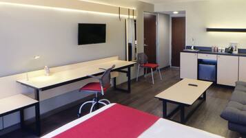 A modern hotel room with a bed, a desk, a chair, a sofa, a TV, and a kitchenette.