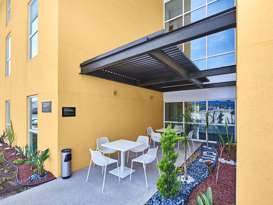 An outdoor seating area with white tables and chairs, surrounded by a yellow building facade and a glass window.