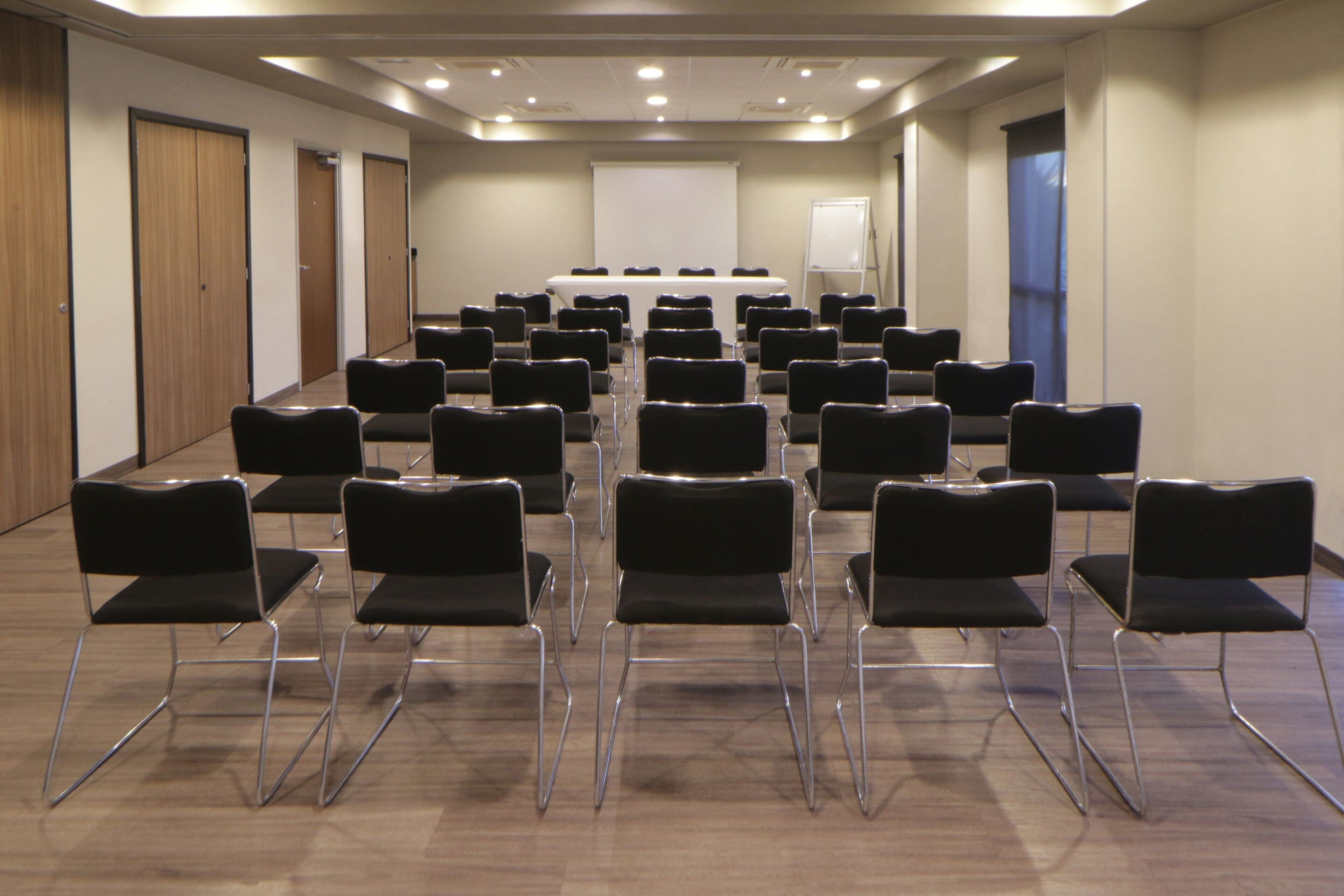 Une salle de conférence avec des rangées de chaises disposées face à un tableau blanc, des portes en bois et un tableau blanc sur le mur de droite.