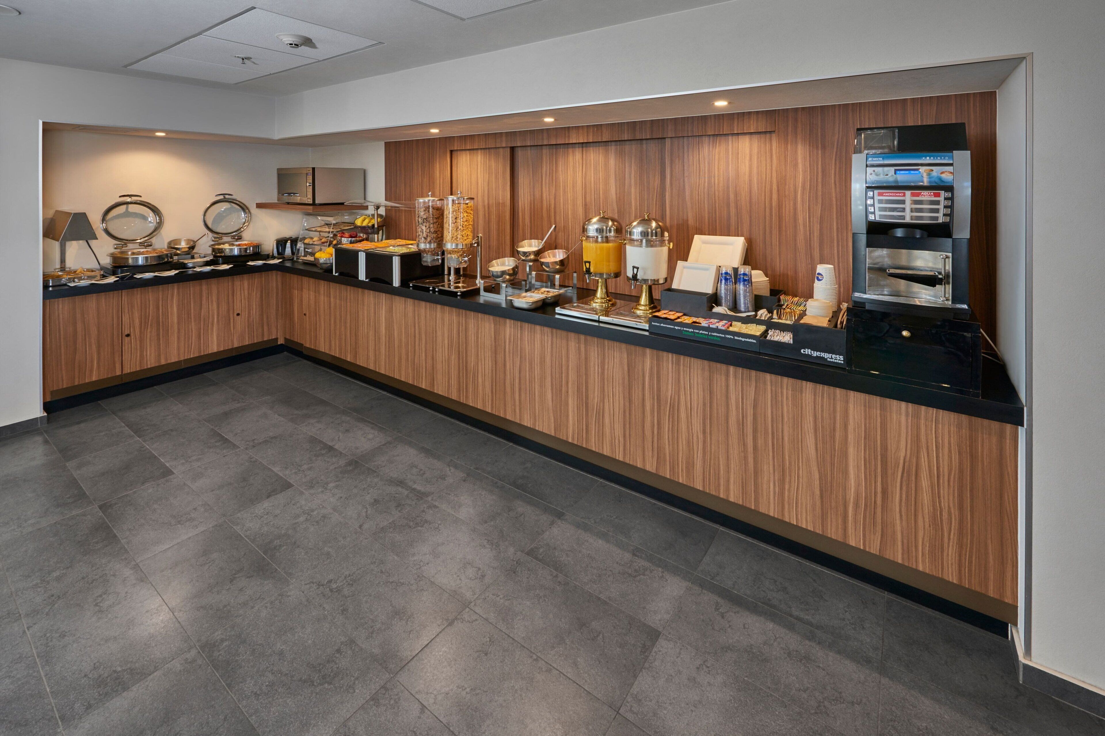 A modern hotel breakfast area with a wooden counter, self-service beverage station, and a microwave.