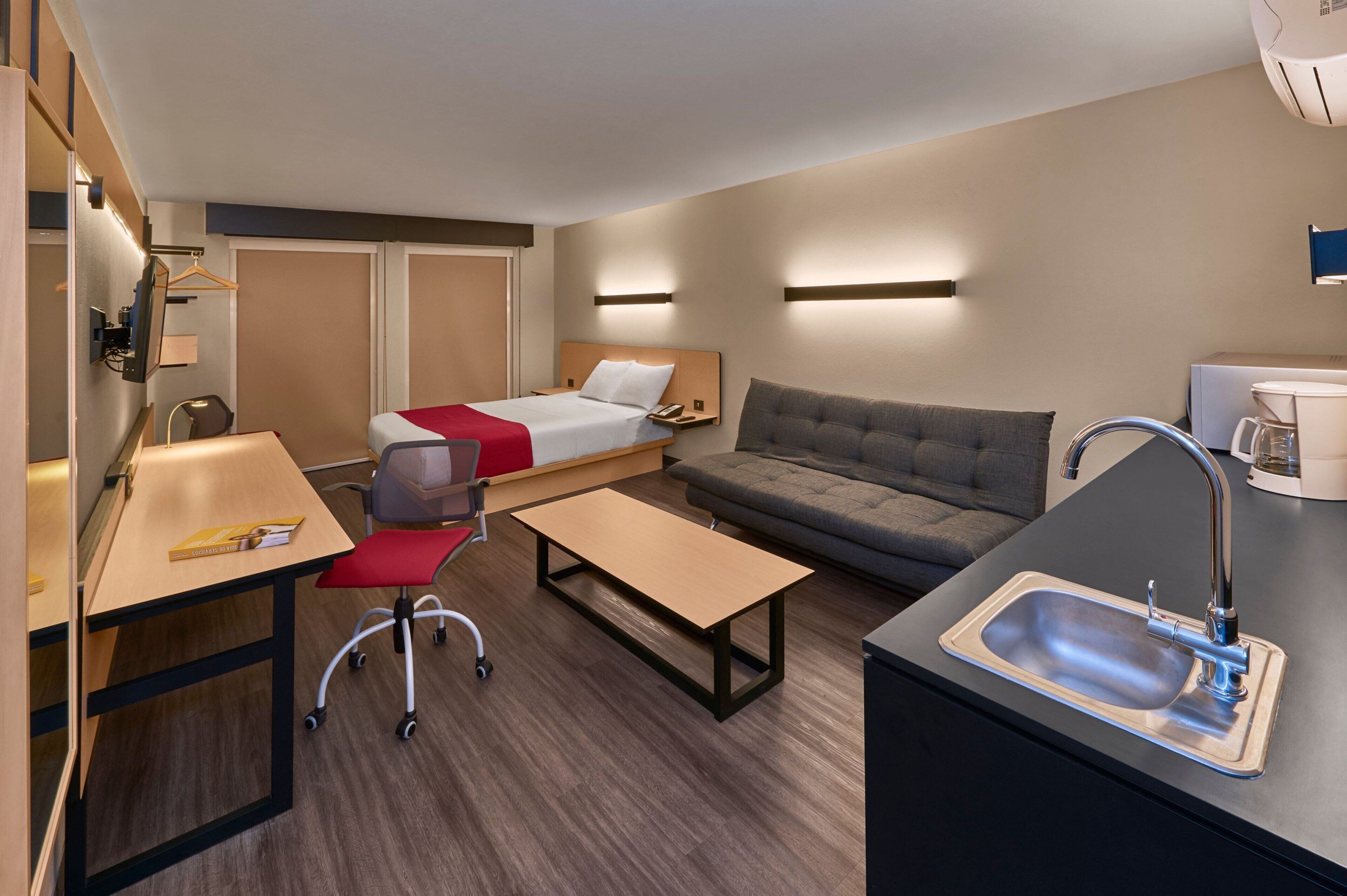 Habitación de hotel pequeña con cama, sofá, escritorio con silla, mesita, lavamanos y espejo.