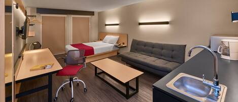 Habitación de hotel pequeña con cama, sofá, escritorio con silla, mesita, lavamanos y espejo.