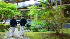 Un jardin japonais traditionnel, avec des allées pavées de pierres, une végétation luxuriante et un bâtiment en bois surmonté d’un toit en tuiles.