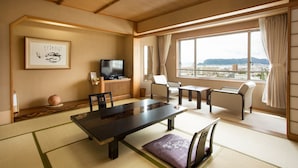 Une pièce de style japonais traditionnel, avec un sol recouvert de tatamis, une table basse, des chaises, un téléviseur et une vue sur le paysage urbain à travers de grandes fenêtres.