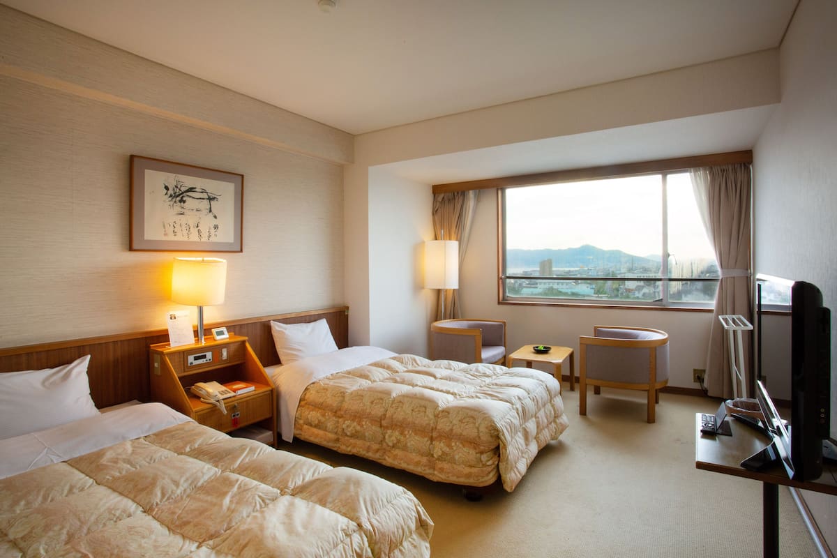 Une chambre d’hôtel avec deux lits, un bureau avec un ordinateur, une chaise, une lampe et une fenêtre donnant sur la ville.