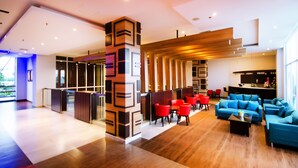 Un moderno vestíbulo de hotel con un mostrador de recepción de madera, zonas de estar en rojo y azul, y un amplio ventanal que proporciona luz natural.