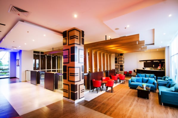 A modern hotel lobby with a wooden reception desk, red and blue seating areas, and a large window providing natural light.