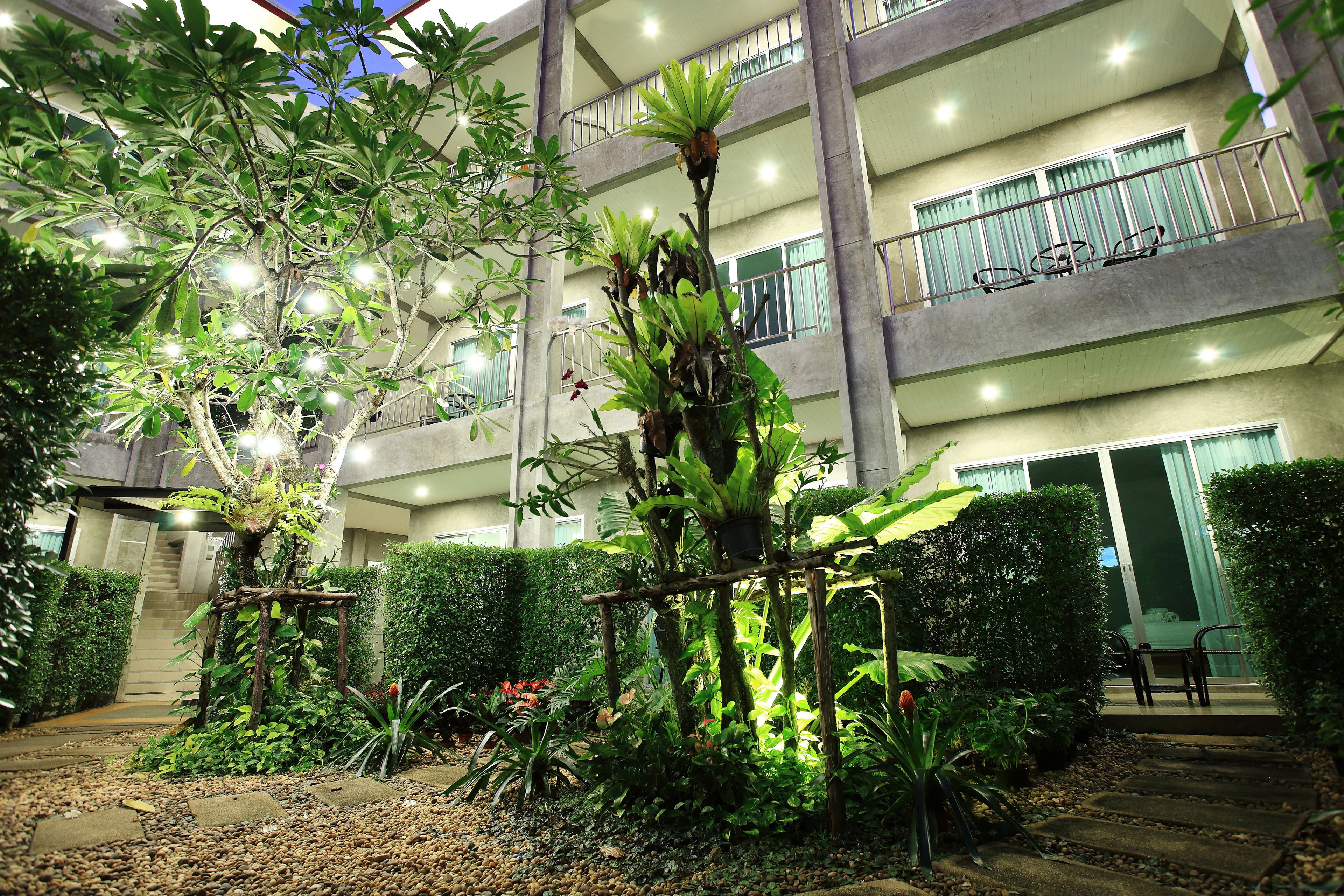 A multi-story building with balconies, a garden area with various plants, and a pathway leading to the entrance.