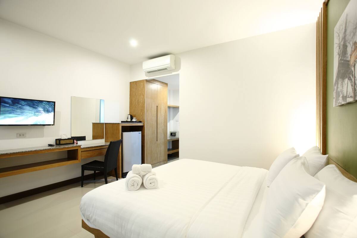 A hotel room with a bed, a desk, a chair, a TV, and an air conditioning unit.