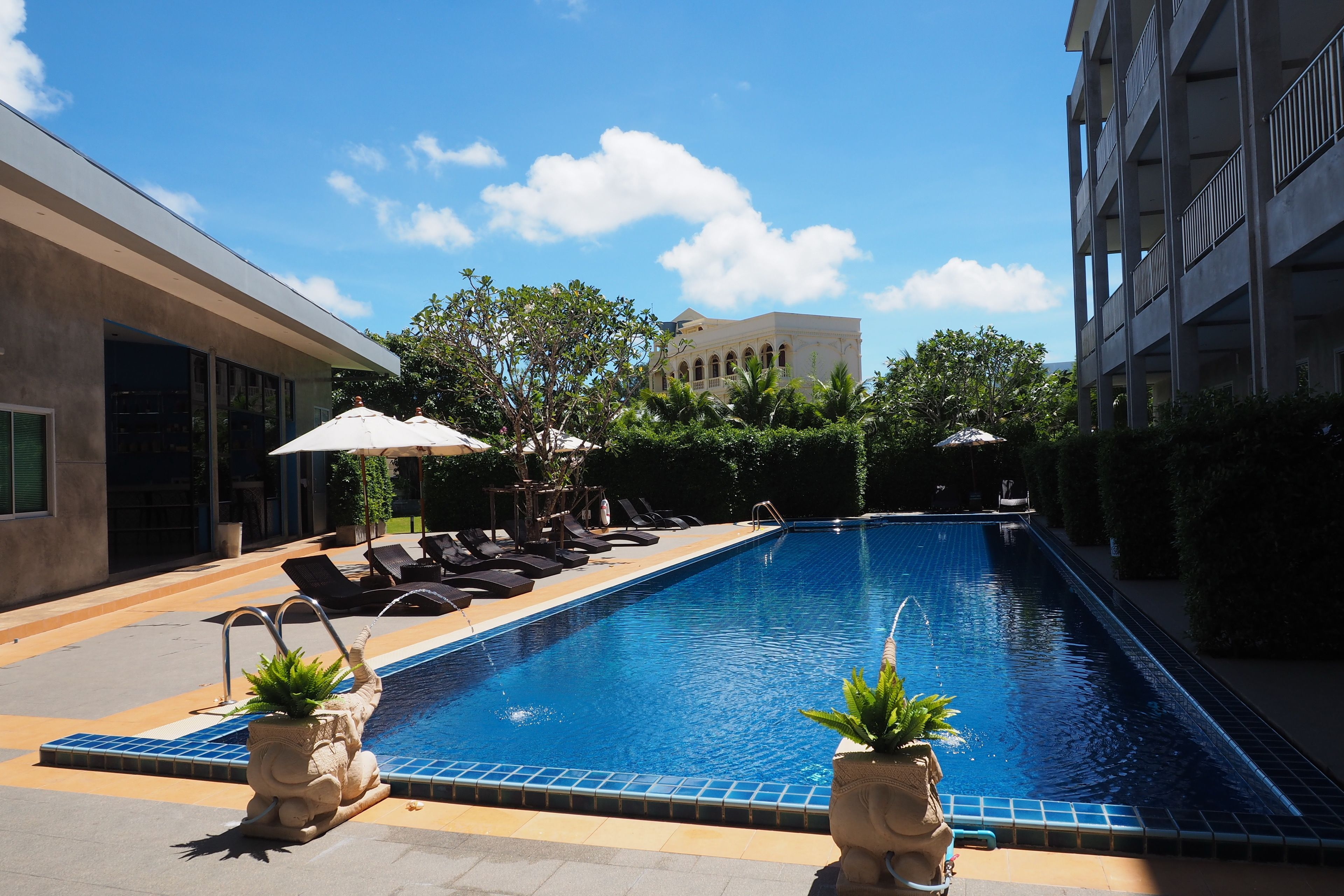 A swimming pool with lounge chairs and umbrellas, surrounded by greenery and a building in the background.