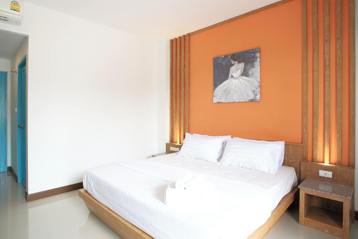 A neatly made bed with white linens in a room with an orange accent wall and a painting of a woman in a white dress.