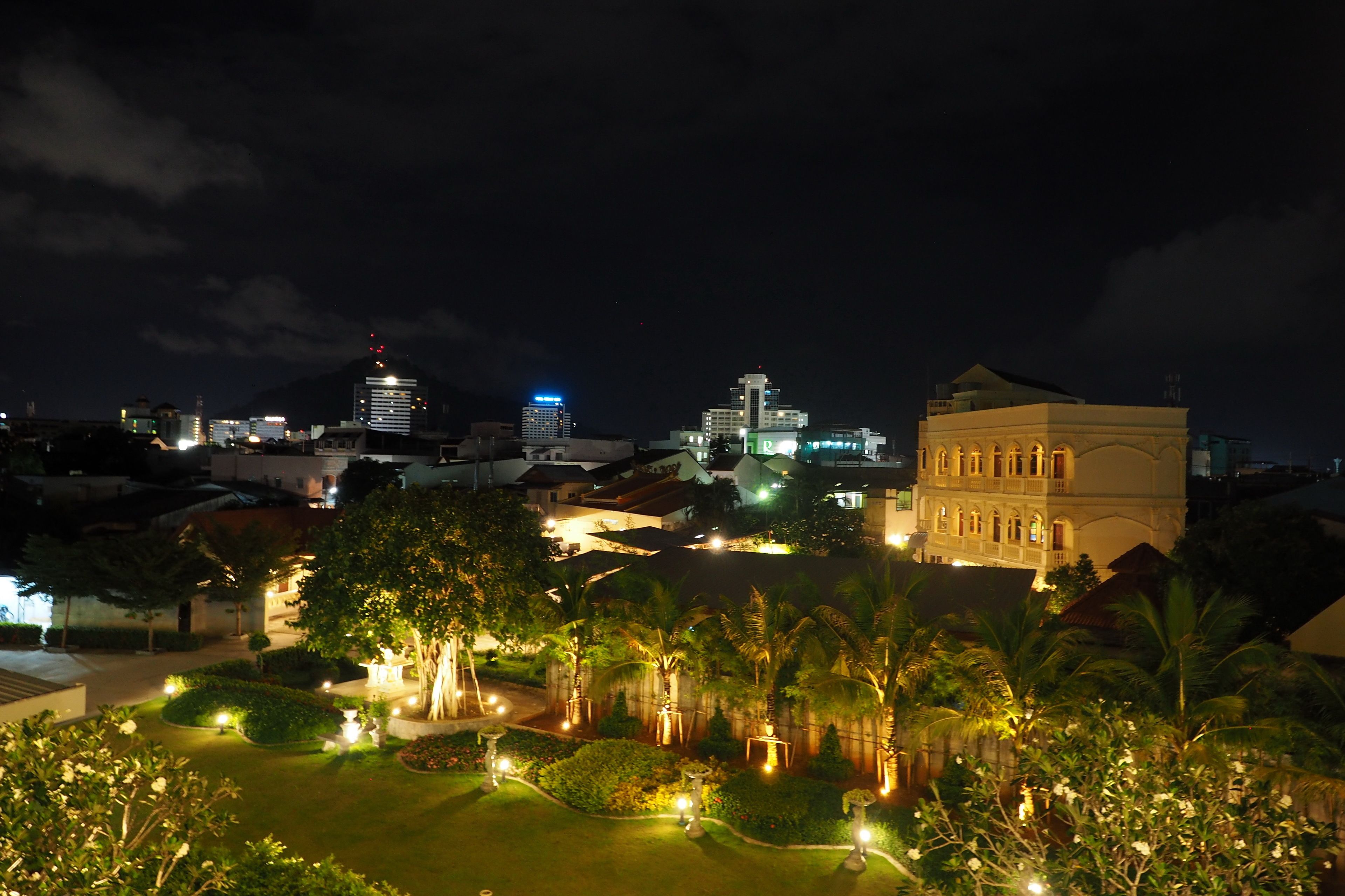 A nighttime cityscape with illuminated buildings, a well-lit garden area with pathways, and a prominent building with classical architecture.