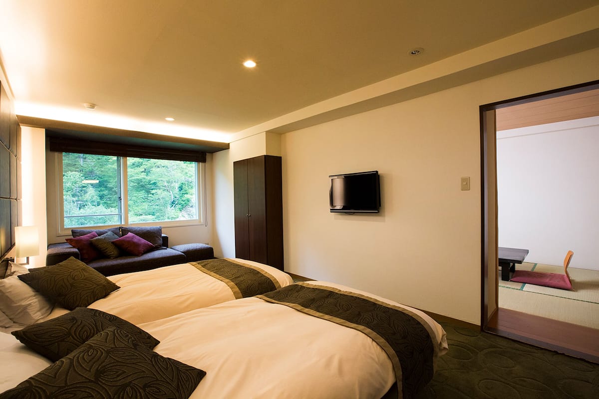 Ein Hotelzimmer mit zwei Betten, einem an der Wand befestigten Fernseher und einem Fenster mit Blick auf Grünflächen.