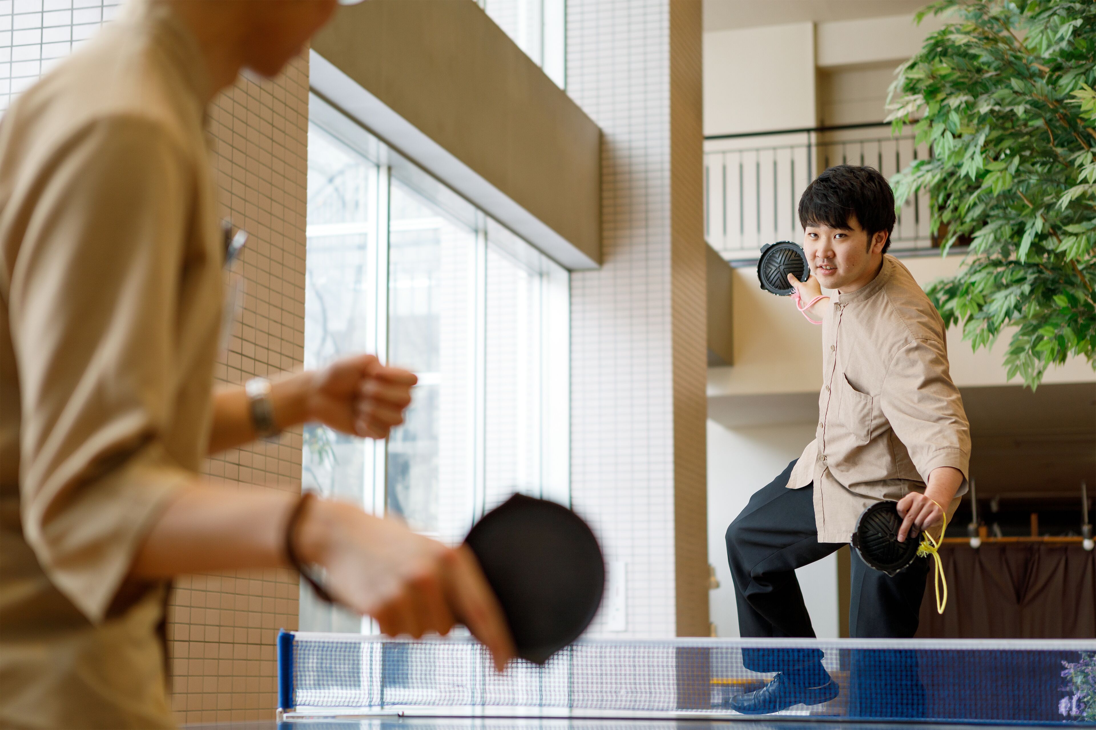Una mesa de ping pong en un espacio interior moderno, con grandes ventanales y una planta de fondo.