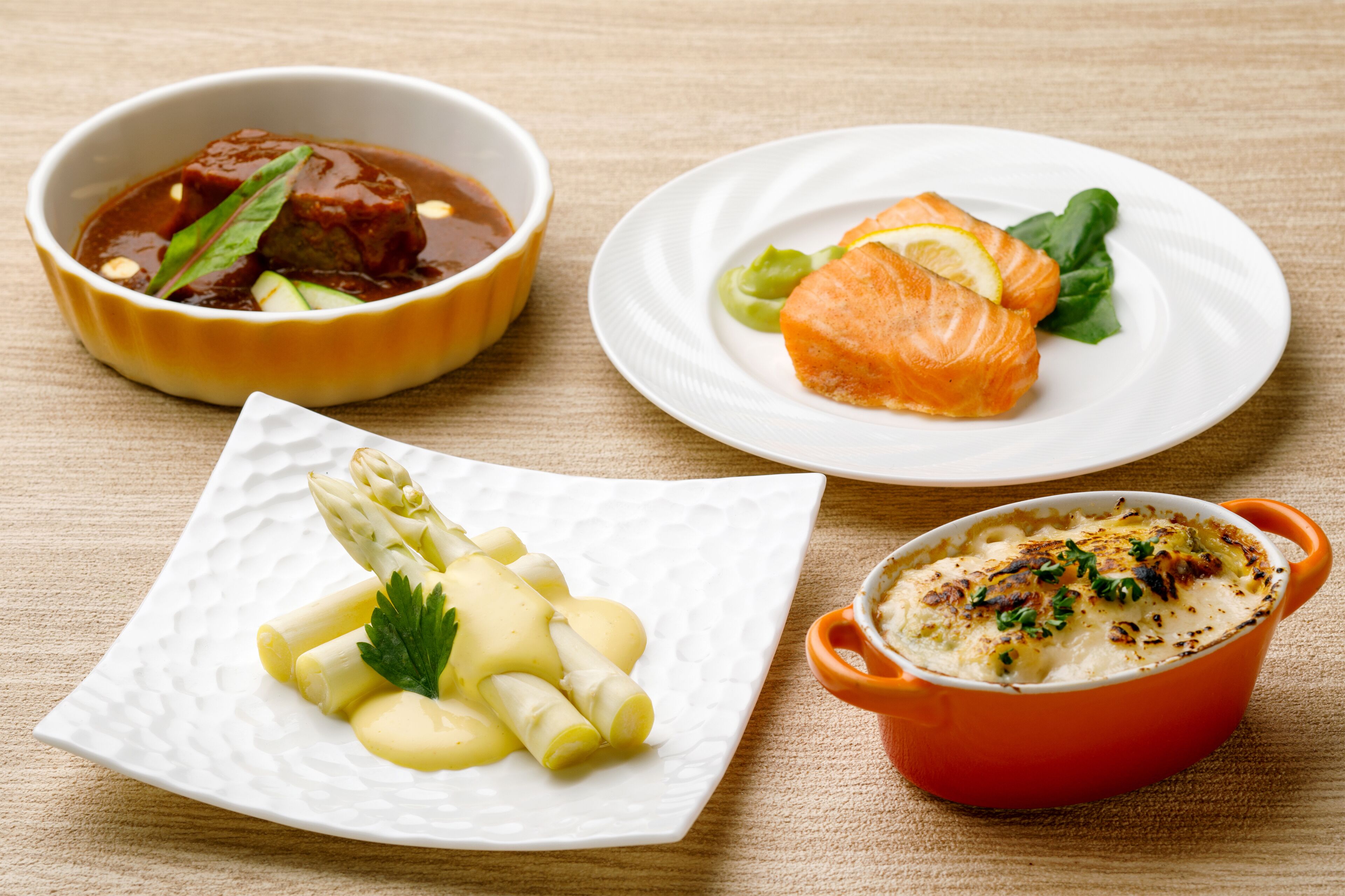 A table with four dishes: a bowl of soup, a plate with salmon and greens, a plate with asparagus and a garnish, and a bowl of baked dish with herbs.