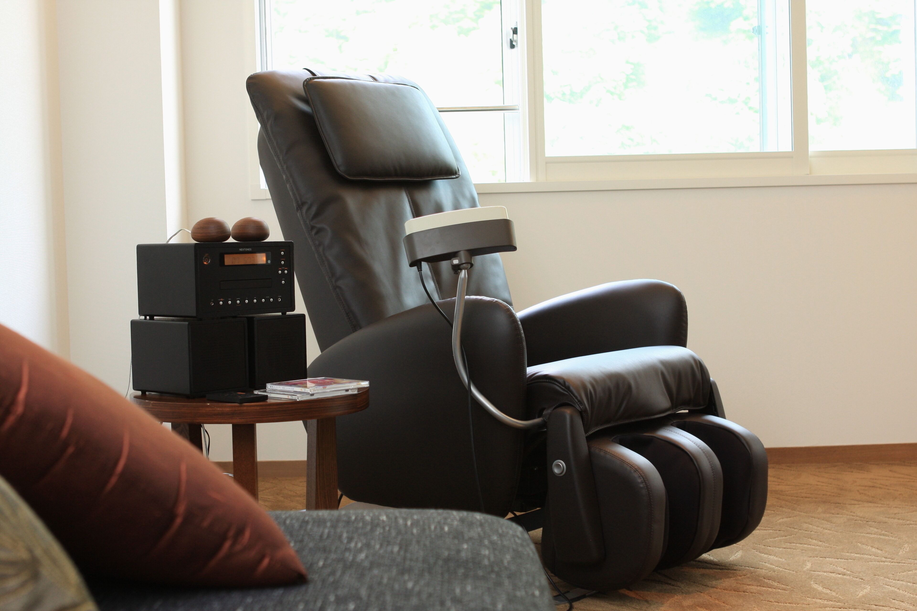 A massage chair with a control panel, a small table with a book and a remote, and a window with a view of trees.