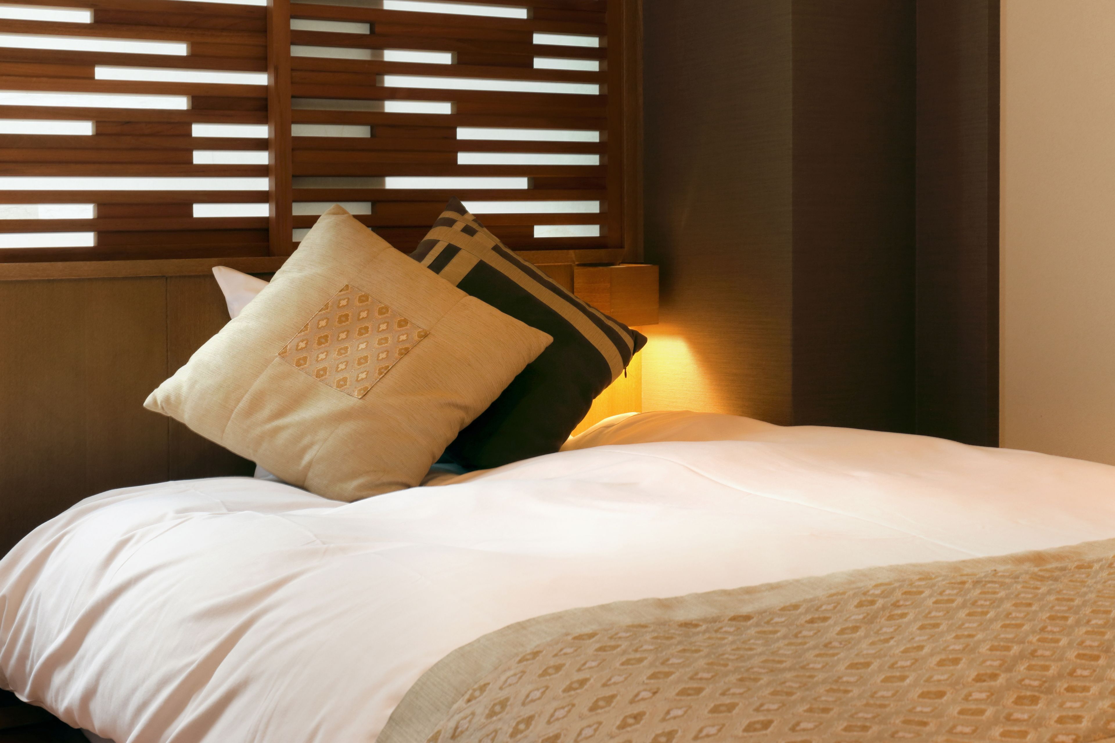 Een opgemaakt bed met witte lakens en twee decoratieve kussens, een houten spijbelkop en een wandlamp.