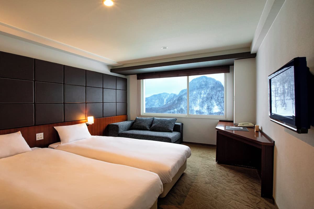Ein Hotelzimmer mit zwei Betten, einem Schlafsofa, einem Schreibtisch und einem großen Fenster mit Blick auf die Berge.