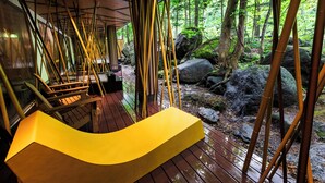 A wooden deck with a yellow bench, surrounded by bamboo poles and a forested area with rocks.