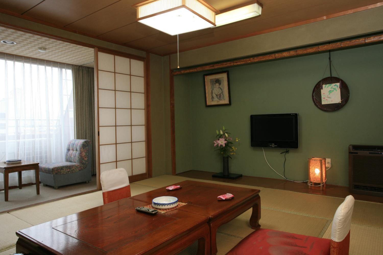 Una camera in stile tradizionale giapponese con pavimento in tatami, un tavolo di legno, una televisione e una piccola scrivania con una lampada.