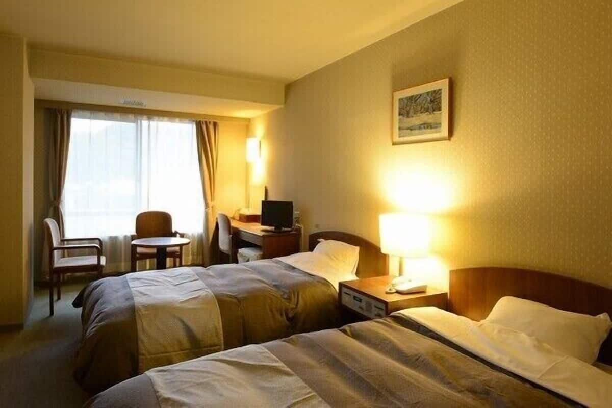 Una habitación de hotel con dos camas, un escritorio con computadora, una silla y una ventana con cortinas.