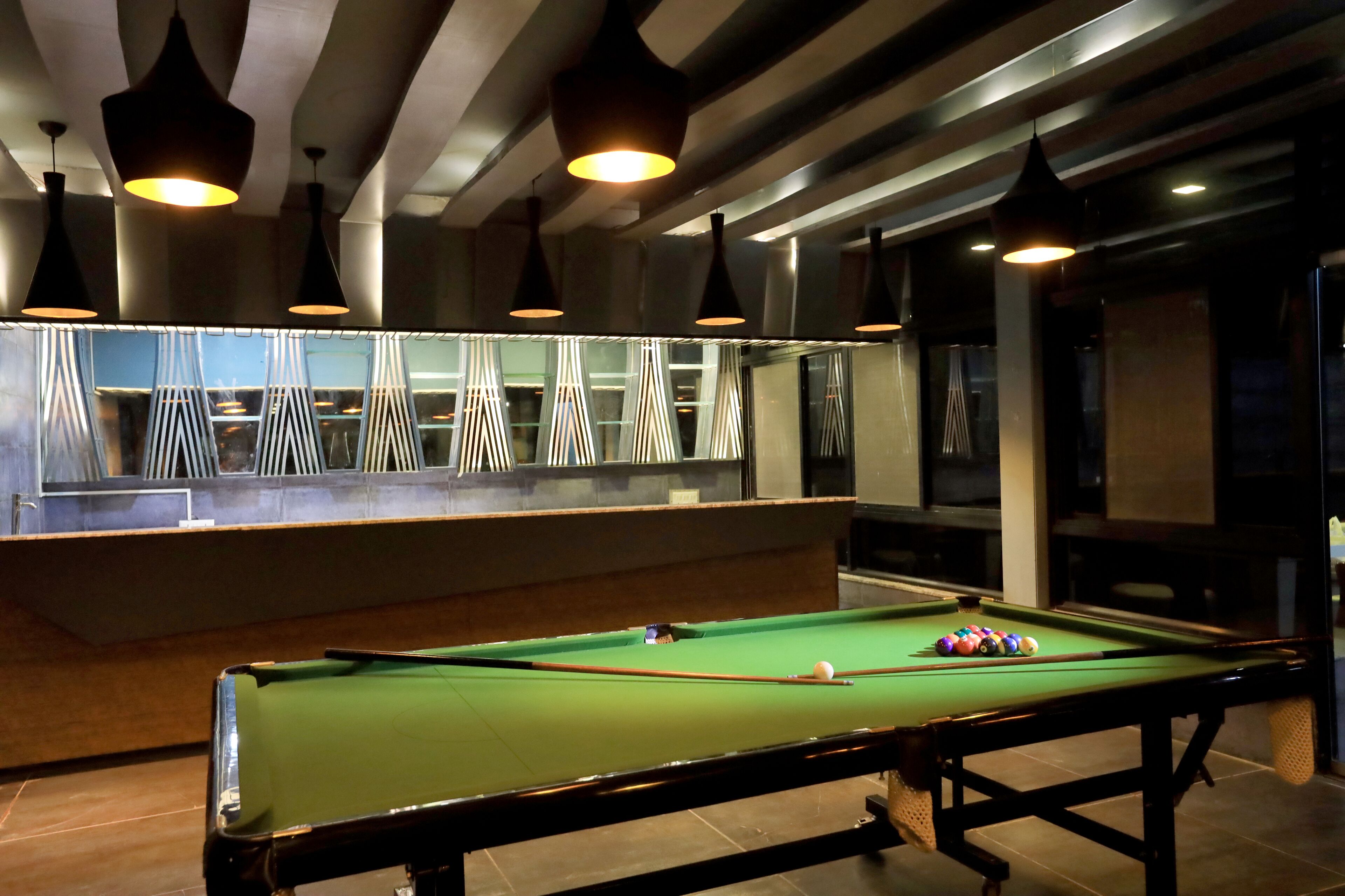 A pool table with balls, overhead lighting, and a bar area with chairs and a counter.