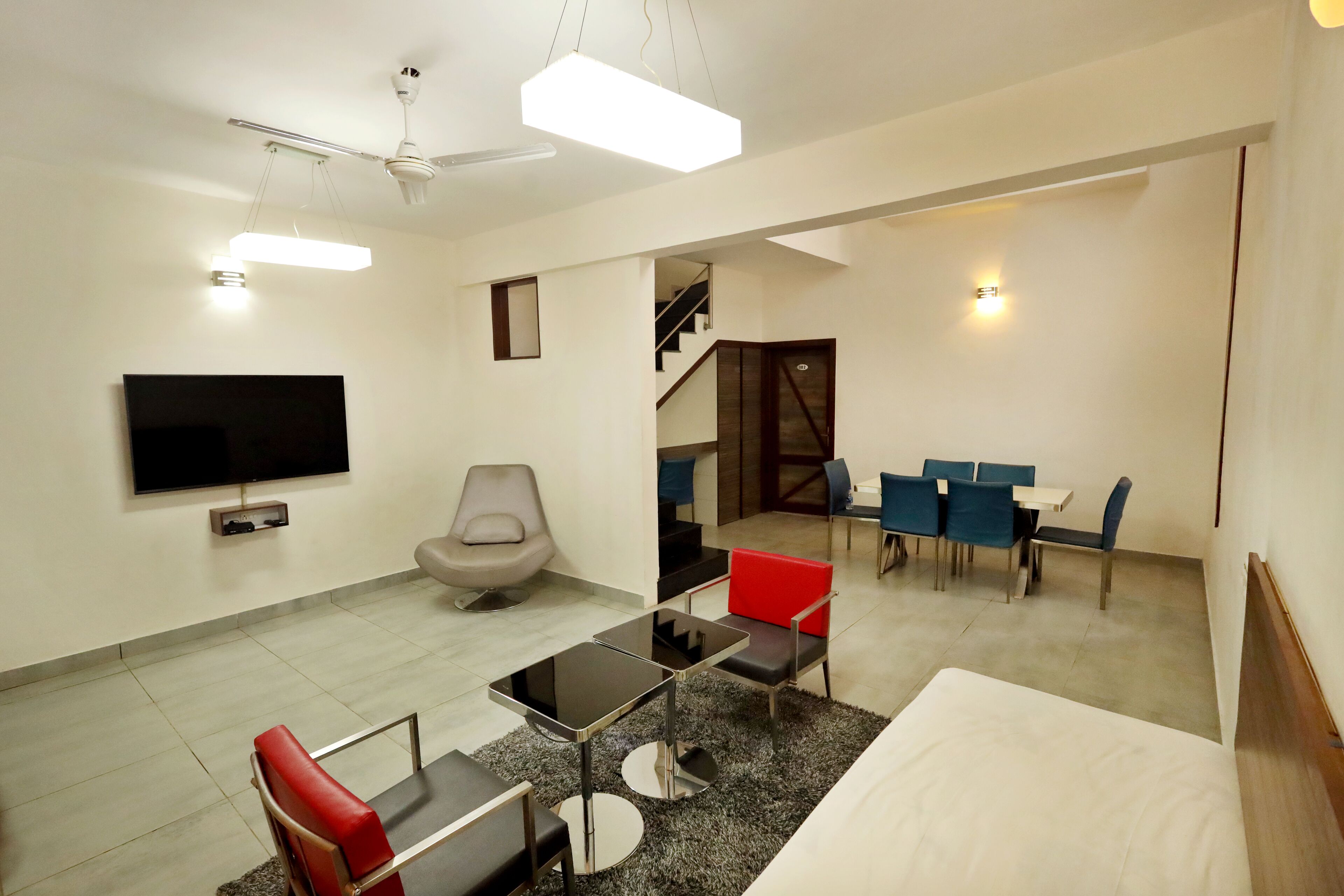 Una camera d'albergo moderna con TV a schermo piatto, un letto, un'area pranzo con sedie blu, una sedia rossa e una scala che conduce a un altro piano.