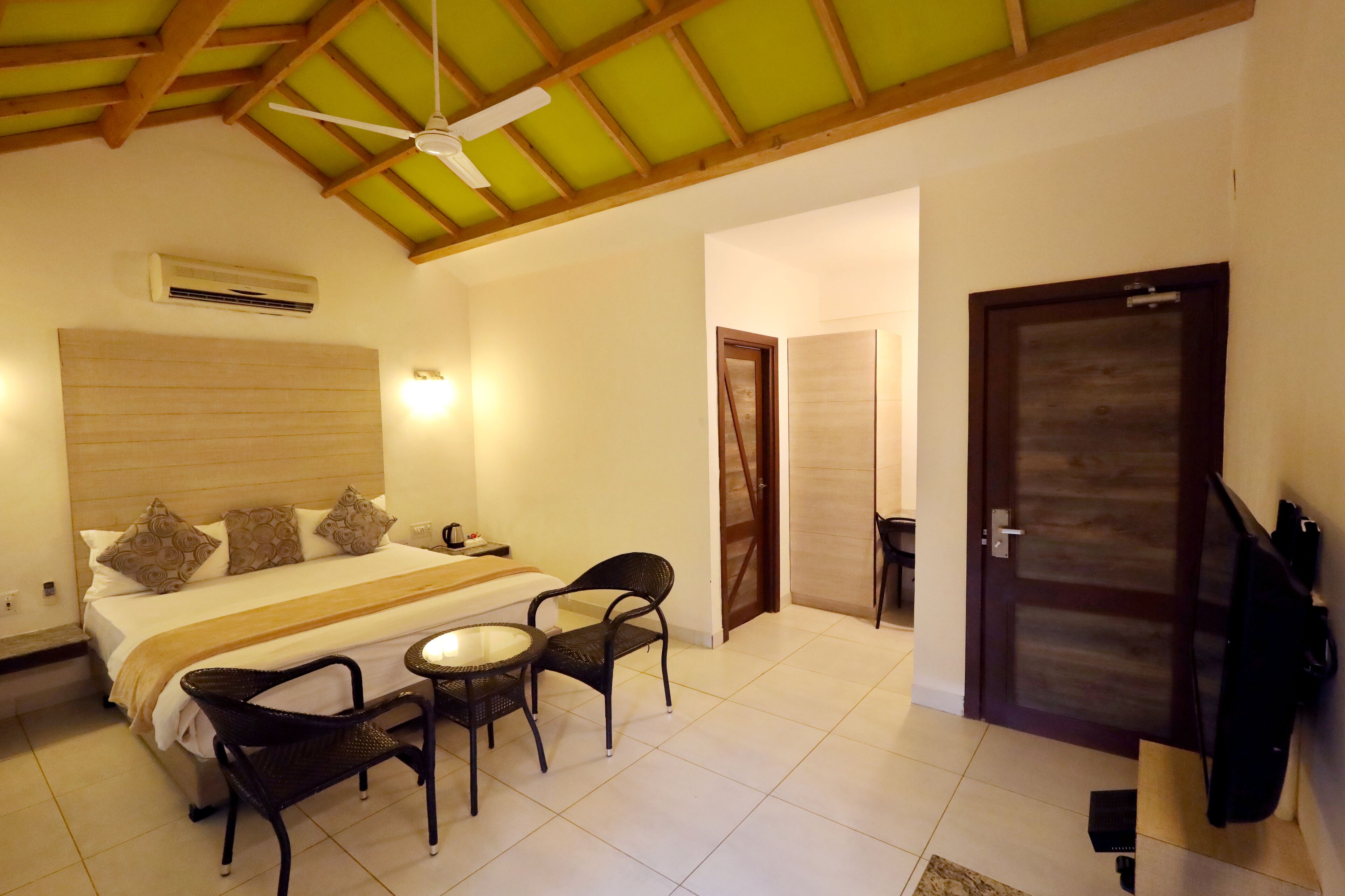 A hotel room with a bed, two chairs, a table, a ceiling fan, an air conditioner, and a TV.