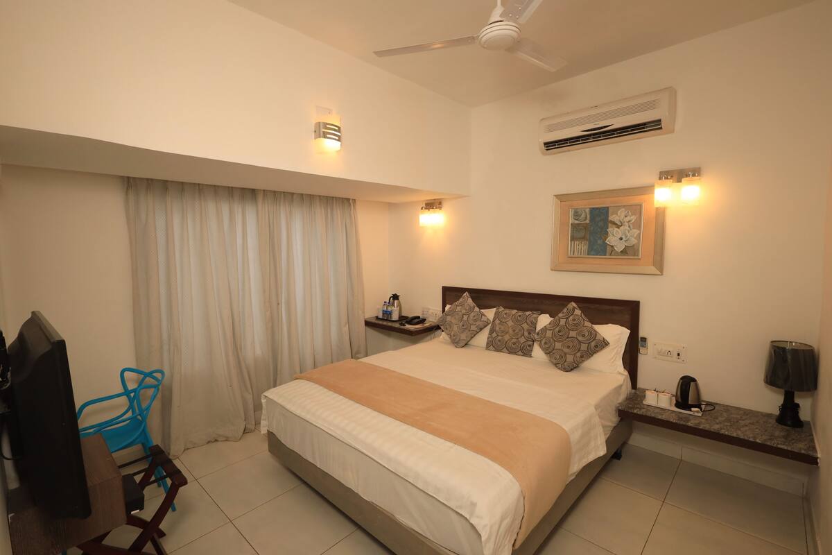 Une chambre d’hôtel comprenant un lit, un bureau avec un ordinateur, une chaise, une fenêtre avec des rideaux et un ventilateur de plafond.