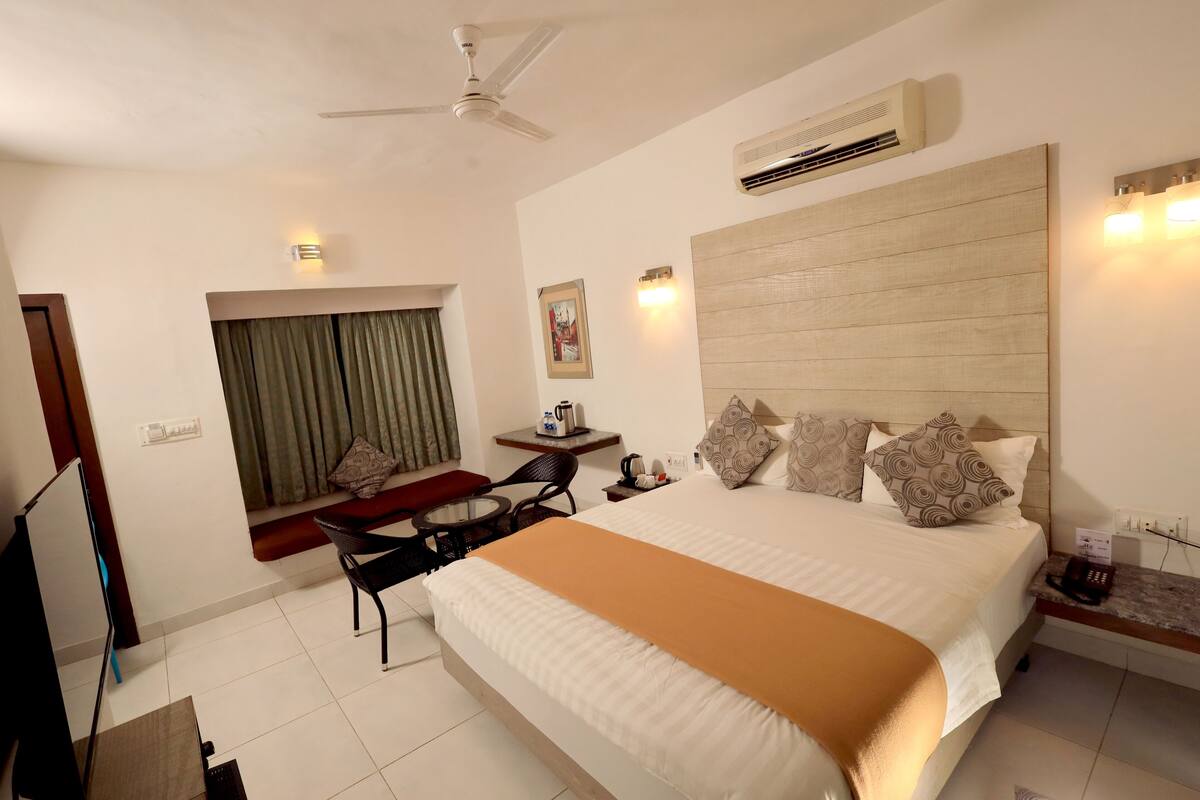 Une chambre d’hôtel avec un grand lit, un ventilateur de plafond, un appareil de climatisation, une petite table avec une chaise, une fenêtre avec des rideaux et un luminaire fixé au mur.