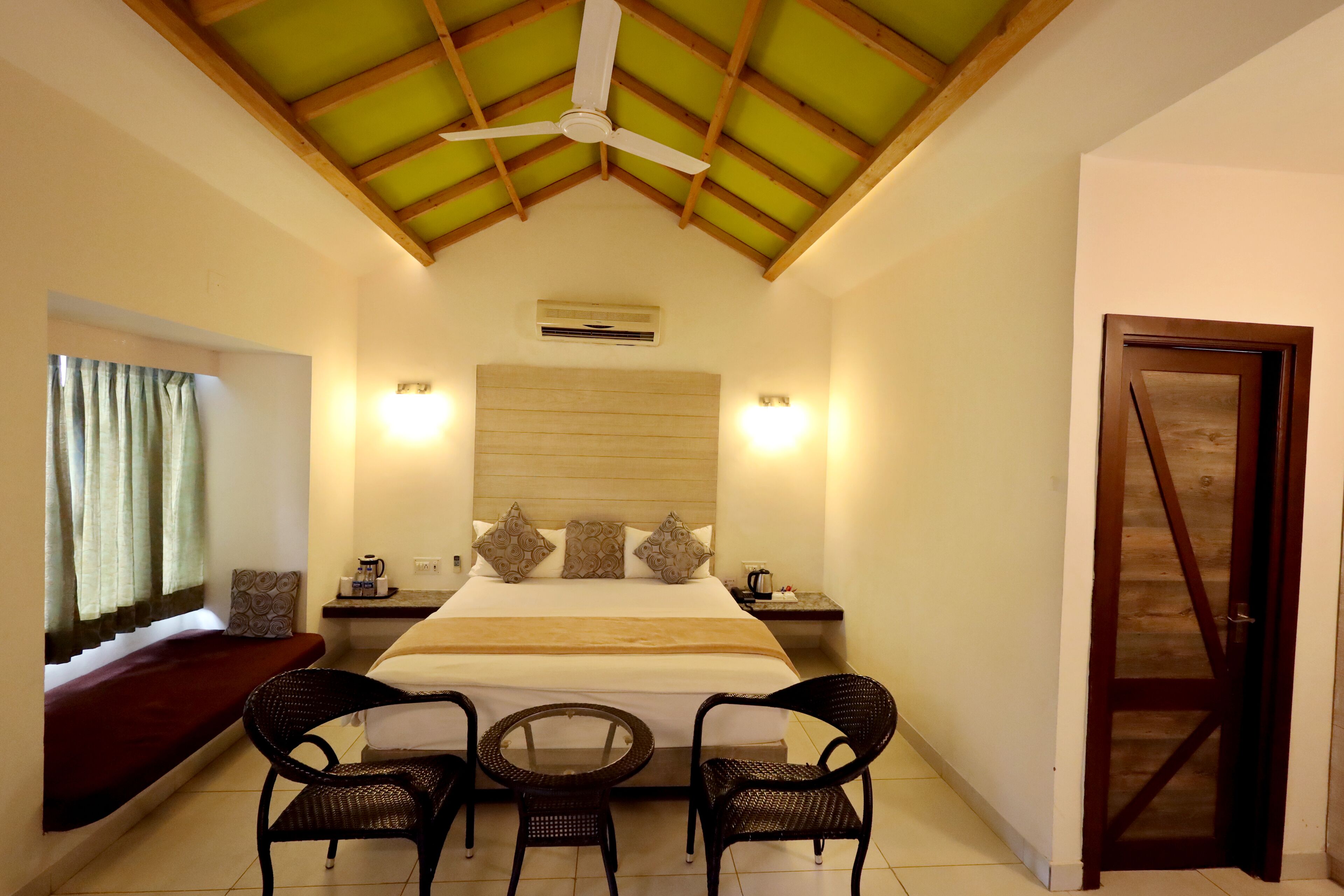 A hotel room with a bed, two chairs, a small table, a nightstand, a window with curtains, a ceiling fan, and a wooden door.