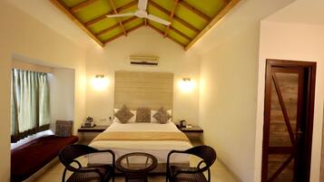 A hotel room with a bed, two chairs, a small table, a nightstand, a window with curtains, a ceiling fan, and a wooden door.