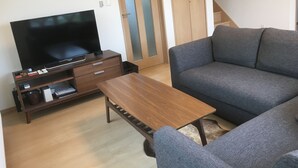 A living room with a flat-screen TV, wooden coffee table, grey sofa, and a wooden wardrobe. - Ume House (Furano)