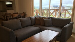 A modern living room with a gray sectional sofa, a wooden coffee table, and a large window with a view of a wooden deck and buildings. - Ume House (Furano)