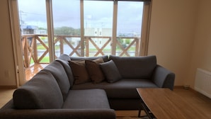 A modern living room with a grey sectional sofa, a wooden coffee table, and a view of a wooden deck through large windows. - Ume House (Furano)