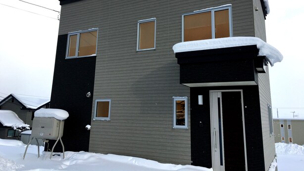 A two-story building with a dark exterior and multiple windows. Snow is visible on the roof and ground. - Ume House (Furano)