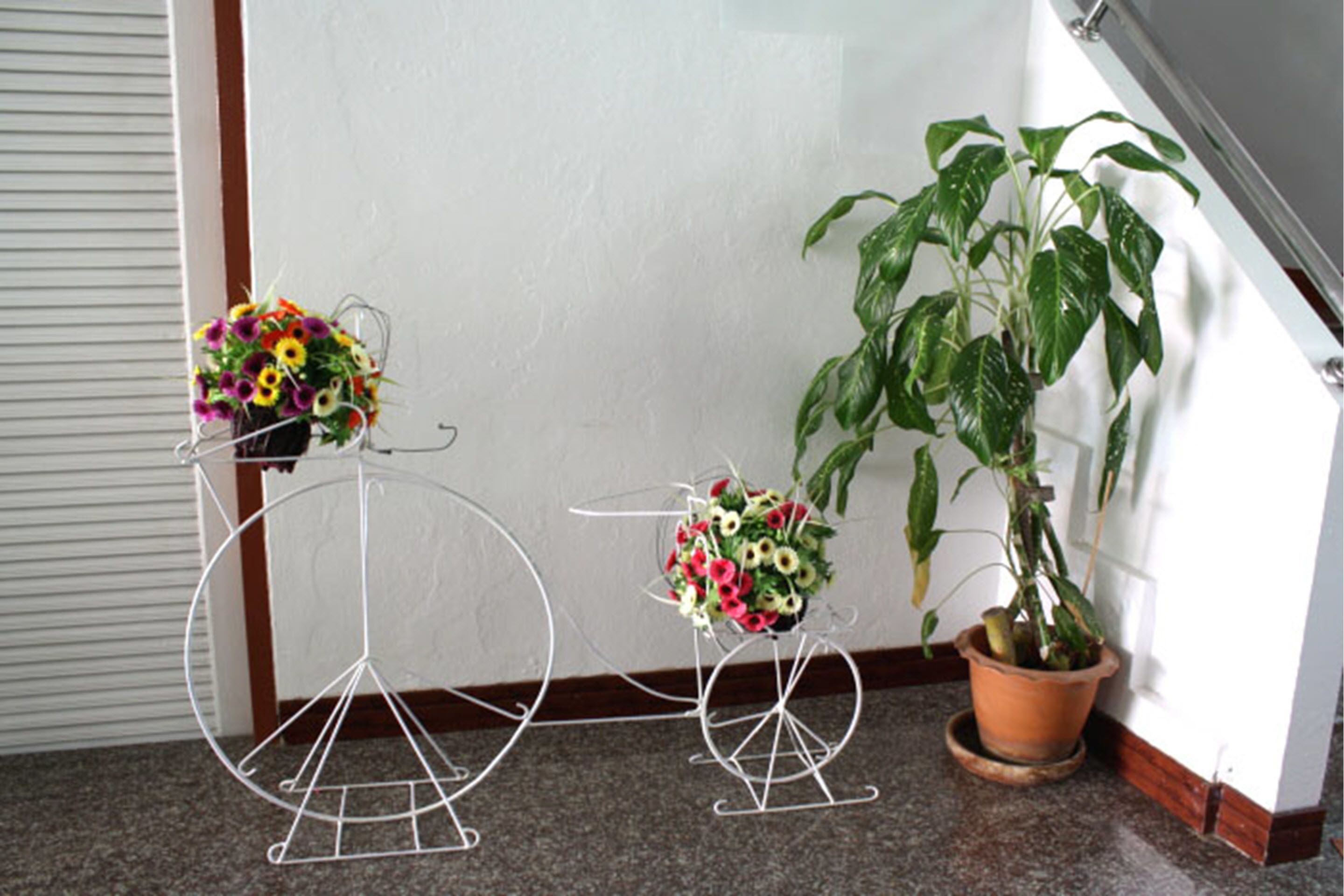 Um canto com um suporte de metal branco exibindo um buquê de flores, uma planta em vaso e uma parede branca.