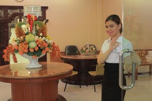 Zona de recepción con un mostrador de madera, un arreglo floral grande y un cartel con el nombre del hotel.