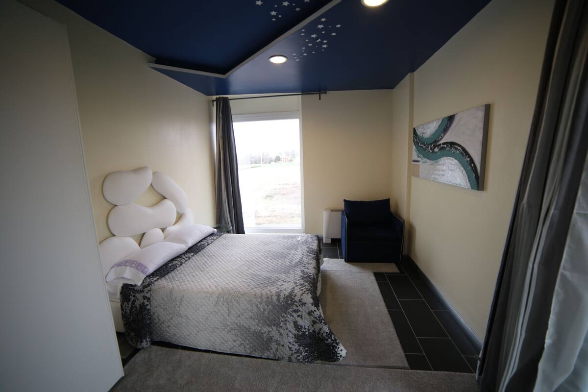A bedroom with a bed, a nightstand, a painting, and a view of the outdoors.
