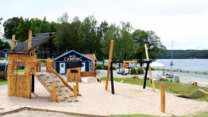 A wooden play structure with a climbing wall and a slide, a small blue building labeled "SKOTTEK CAMPING", and a hammock in a sandy area near a body of water. - Skotteks Gården (Ulricehamn)
