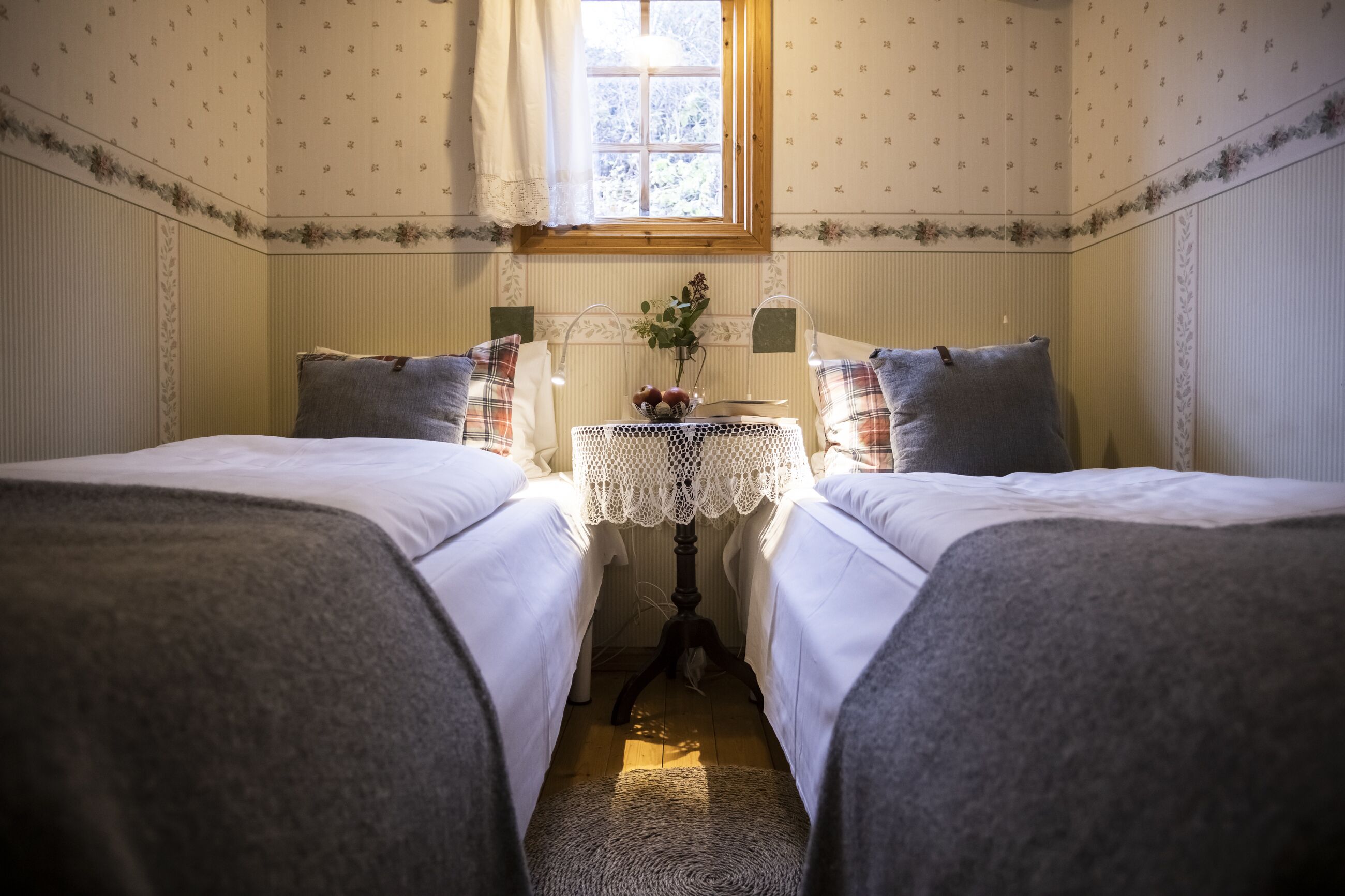 A room with two beds, a small table with a lace tablecloth, and a window with curtains.