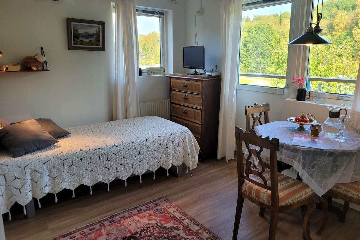 A bedroom with a bed, a dresser, a desk with a computer, a chair, a table with a vase, and a window with a view of trees.