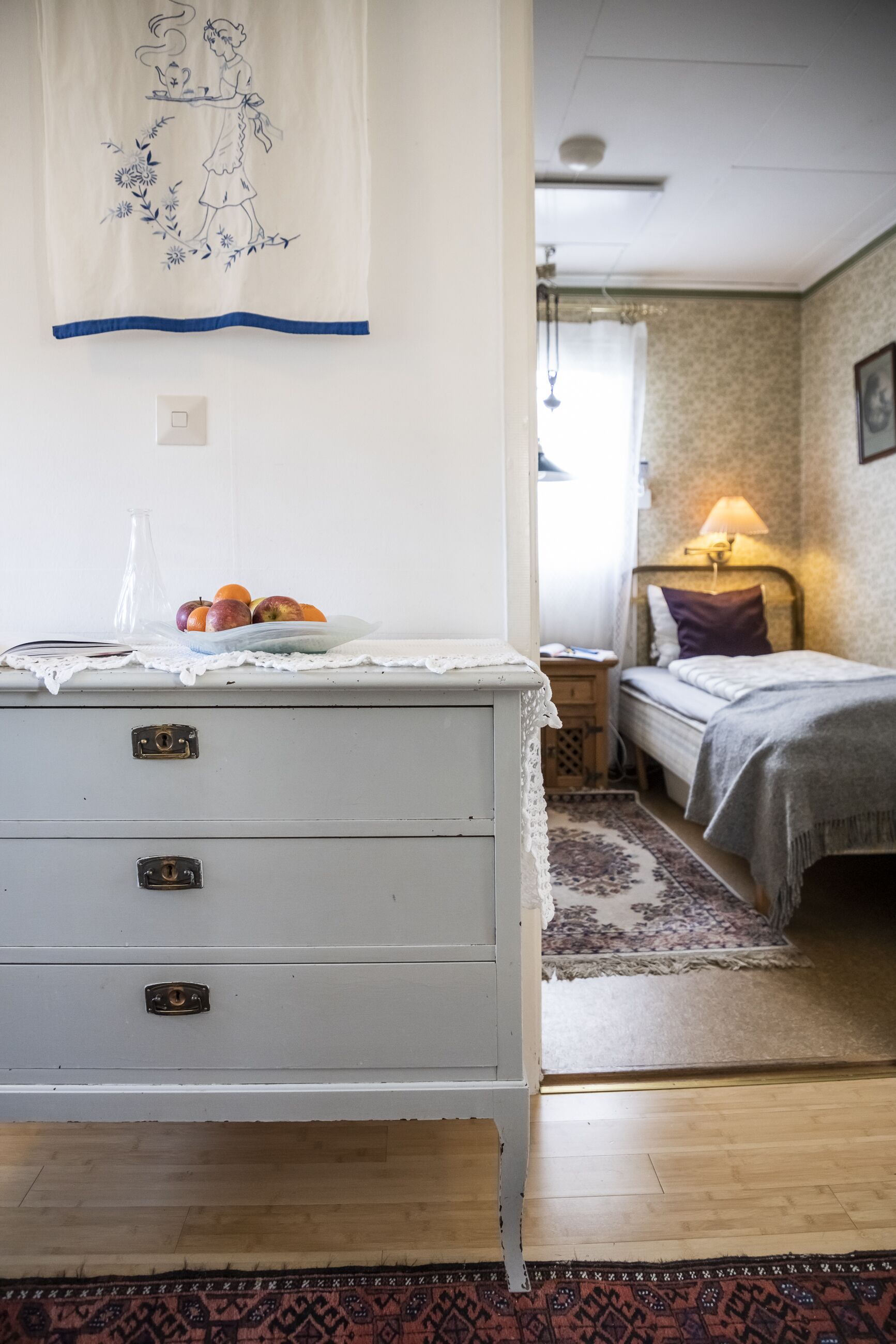 A room with a white dresser, a bed with a patterned bedspread, a nightstand with a lamp, and a wall hanging.