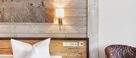 Habitación de hotel con cabecera de madera, lámpara de noche, cama con ropa blanca, mesita de noche, una silla y una obra de arte colgada en la pared.