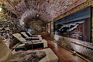 Ein Zimmer mit Steinwänden, einem großen Fenster, Holzboden und mehreren Liegestühlen.