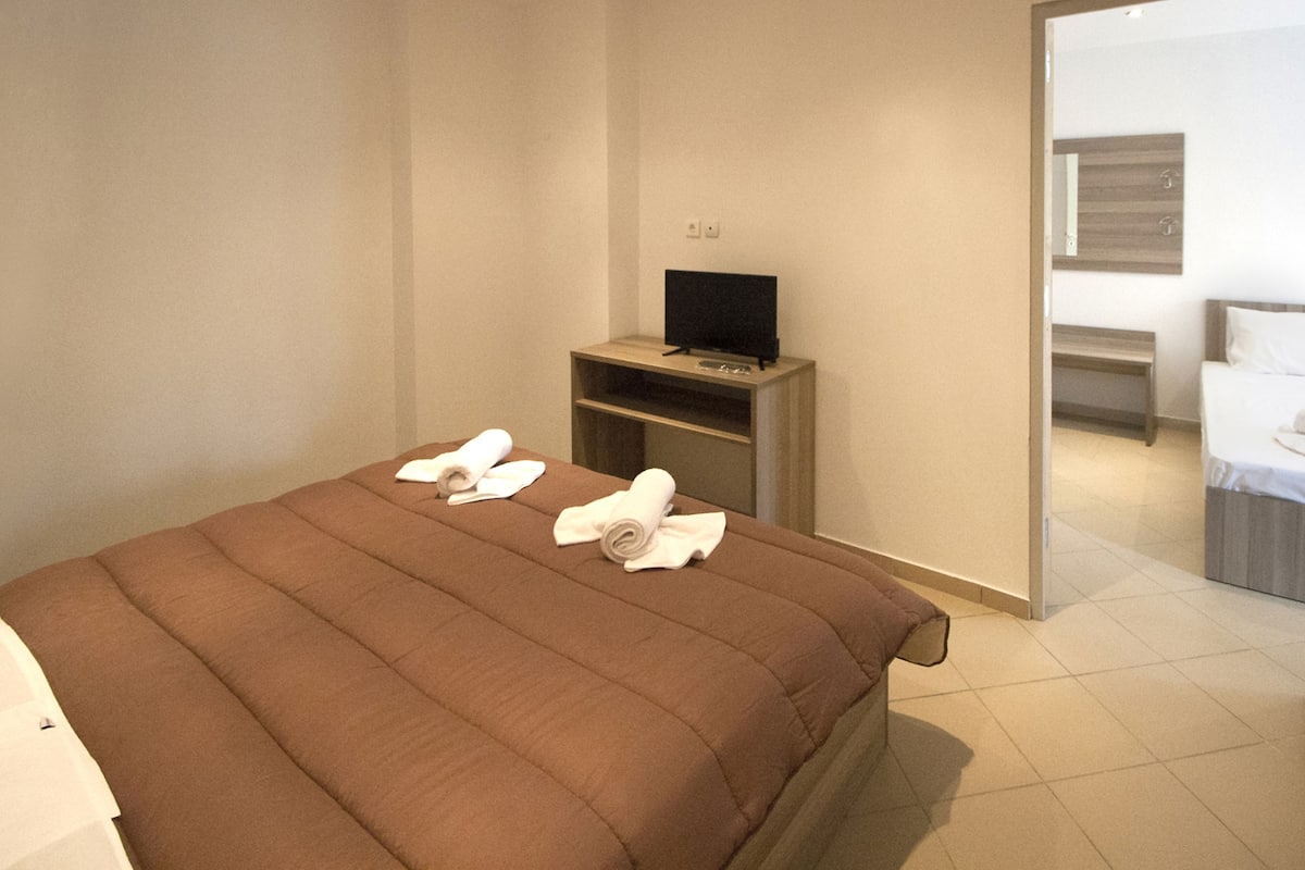 A hotel room with a large bed, a TV on a wooden stand, and a bench in the corner.